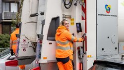Zwei Kolleg:innen auf dem Trittbrett eines Müllwagens
