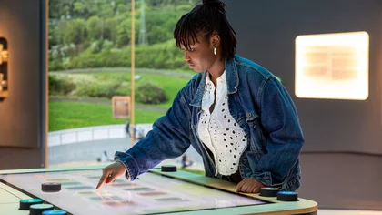 Frau mit Jeansjacke und weißer Bluse steht an einem Touchscreen in der Ausstellung
