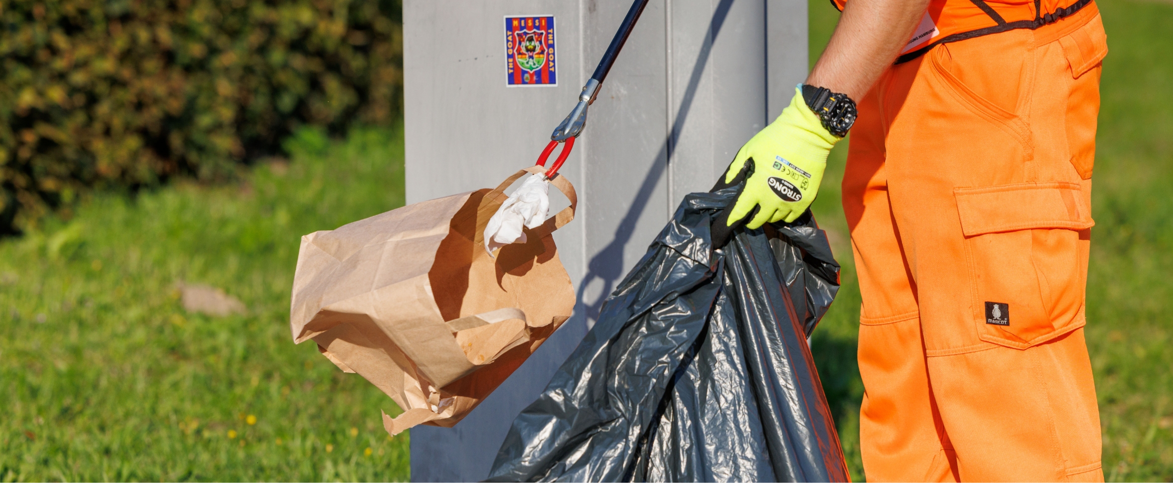 Ein Reiniger in Orange sammelt mit einem Müllgreifer Müll in einen grauen Plastiksack